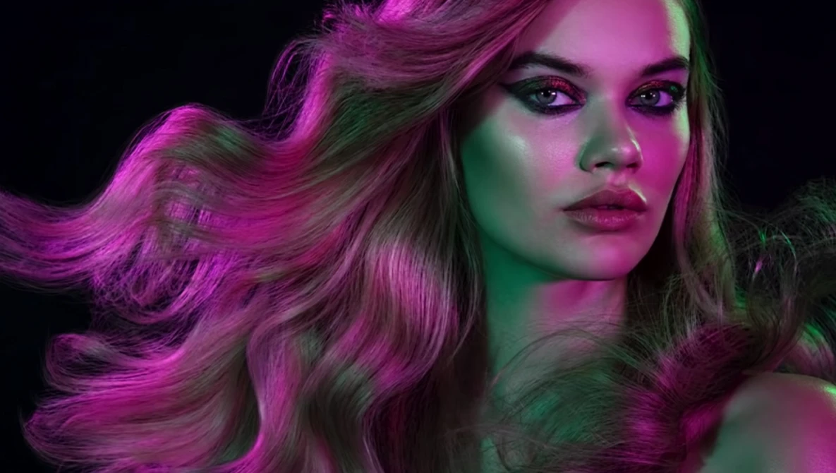 Woman with long wavy hair illuminated by magenta and green gel lighting against a black background.