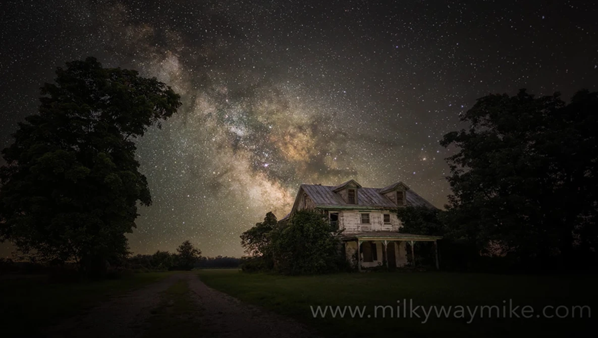 Abandoned two-story farmhouse silhouetted against the Milky Way galaxy stretching across a starry night sky.