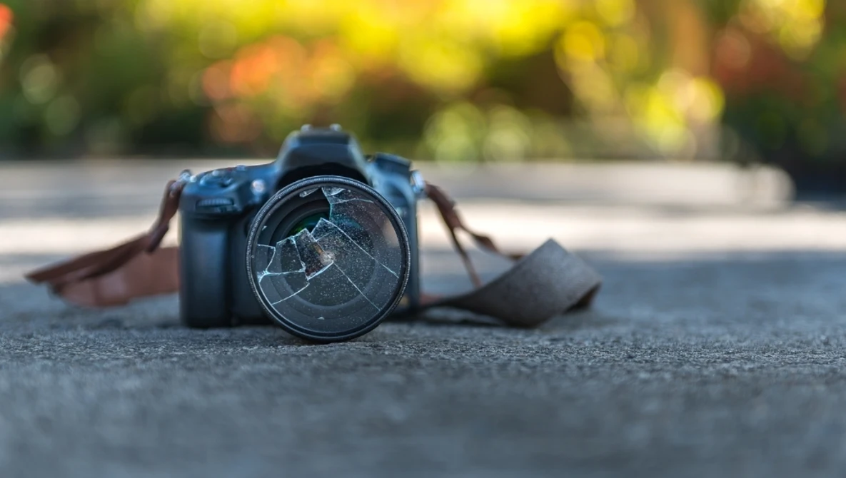 DSLR camera with cracked lens filter lying on pavement with blurred foliage background.
