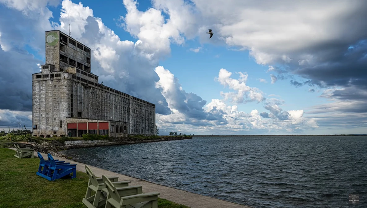 Weathered concrete grain elevator beside calm water with blue Adirondack chair on grassy shore.