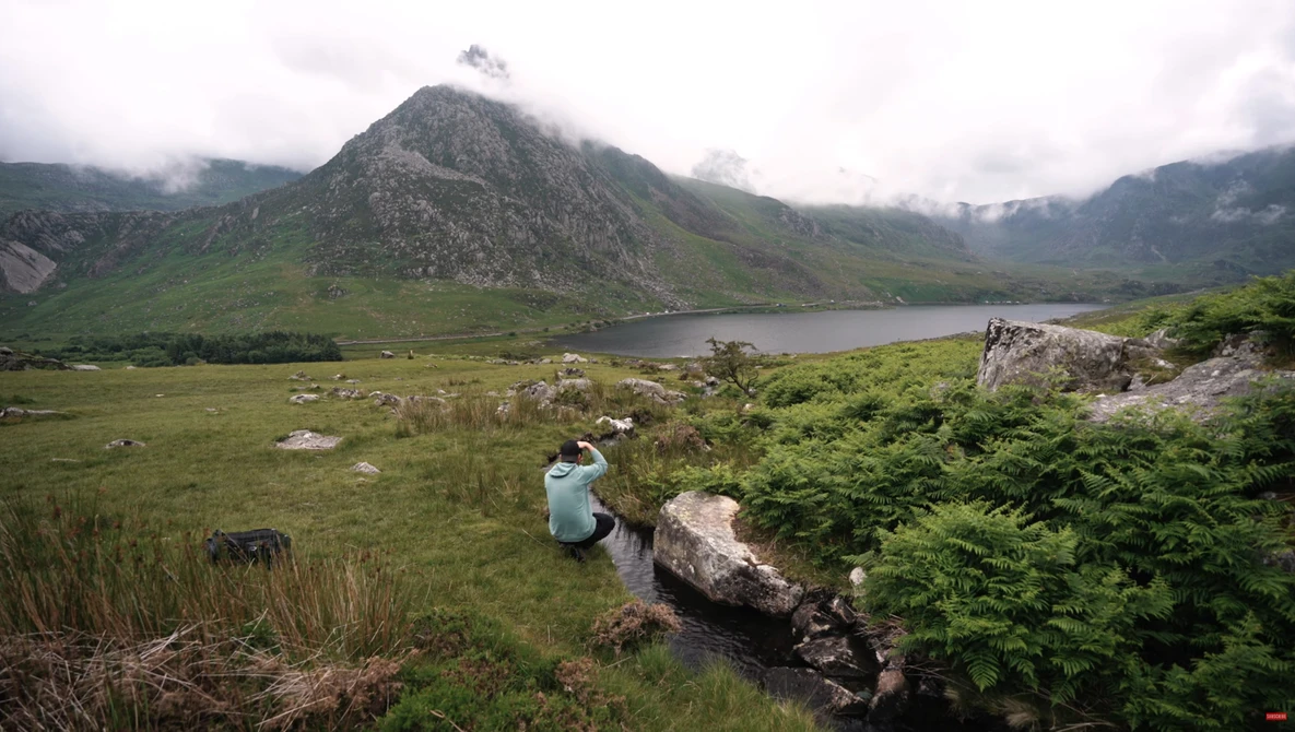 Lone hiker sitting on a rock overlooking a mountain lake surrounded by green valley and peaks.