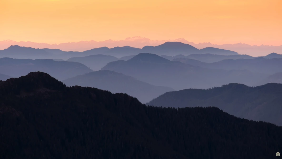 Layered mountain ridges receding into atmospheric haze at sunrise, forested slopes in silhouette.