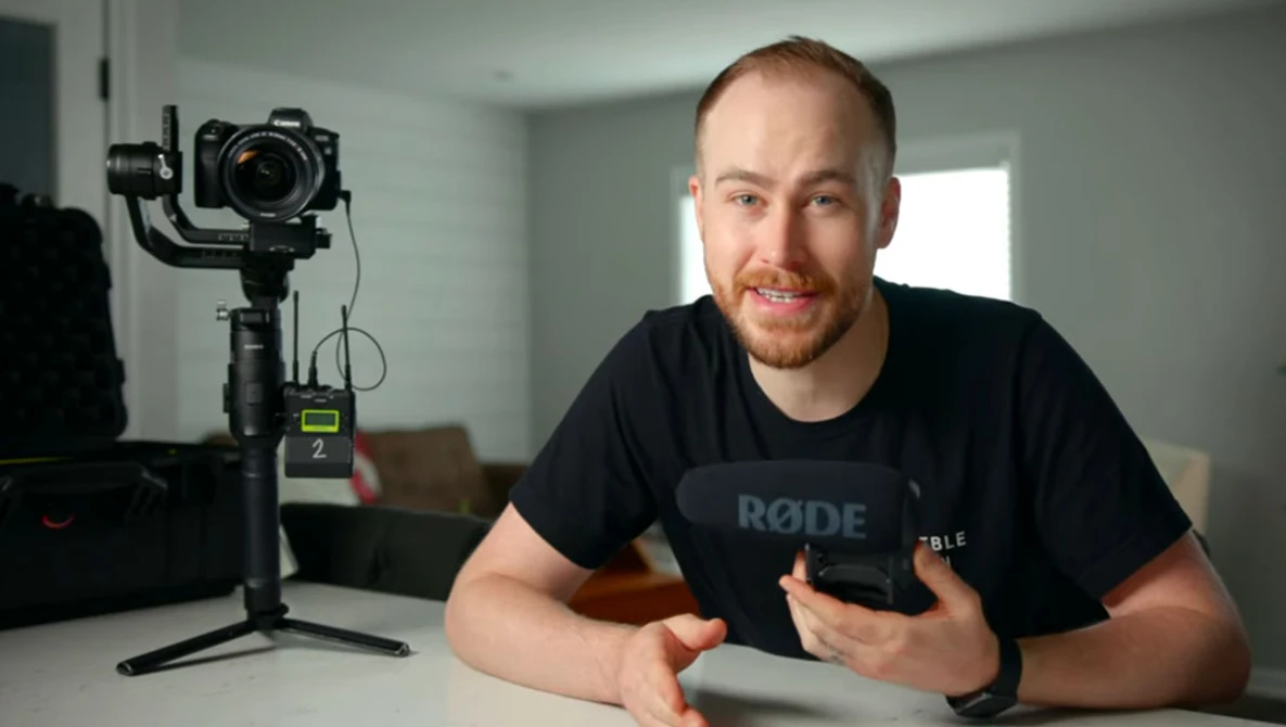 Content creator in black RODE shirt gestures while speaking to camera next to motorized gimbal with mounted DSLR.