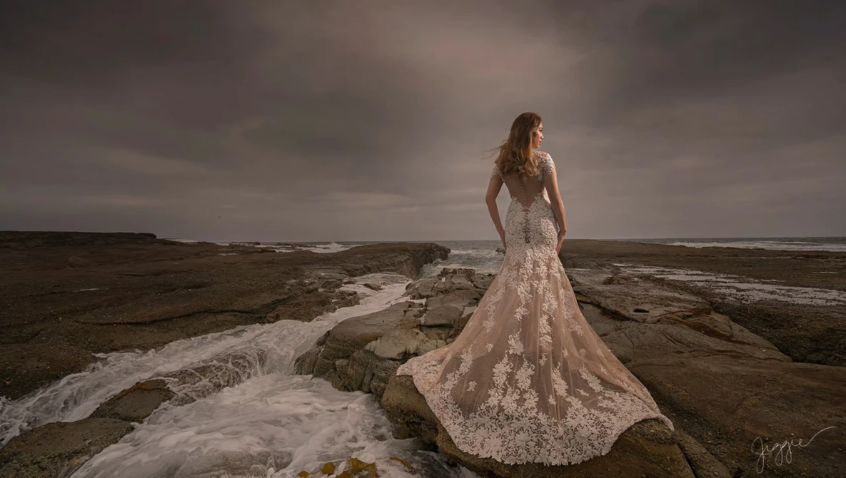 Bride in ornate lace gown standing on rocky coastal outcrop overlooking turbulent ocean.