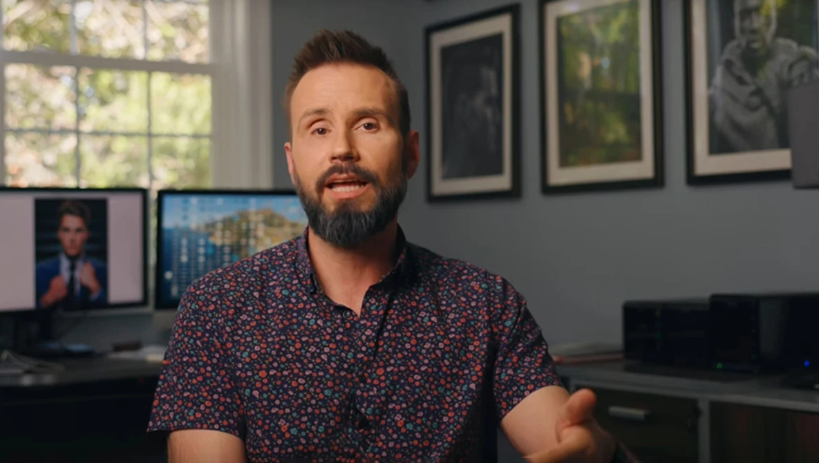 Man with beard wearing patterned short-sleeve shirt in modern office with monitors and framed artwork on walls.