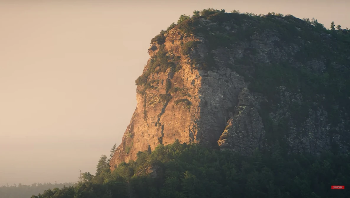 Steep rocky cliff face illuminated by golden hour sunlight with forested summit and misty valley below.