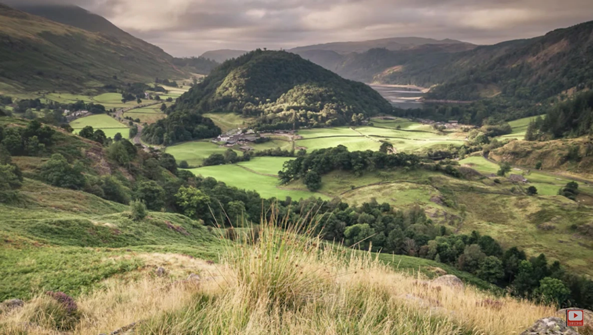Green valley landscape with forested hills and scattered buildings under overcast sky.