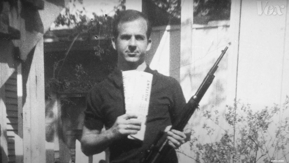 Man in dark cardigan and white shirt holding rifle, posed in front of white building with foliage.