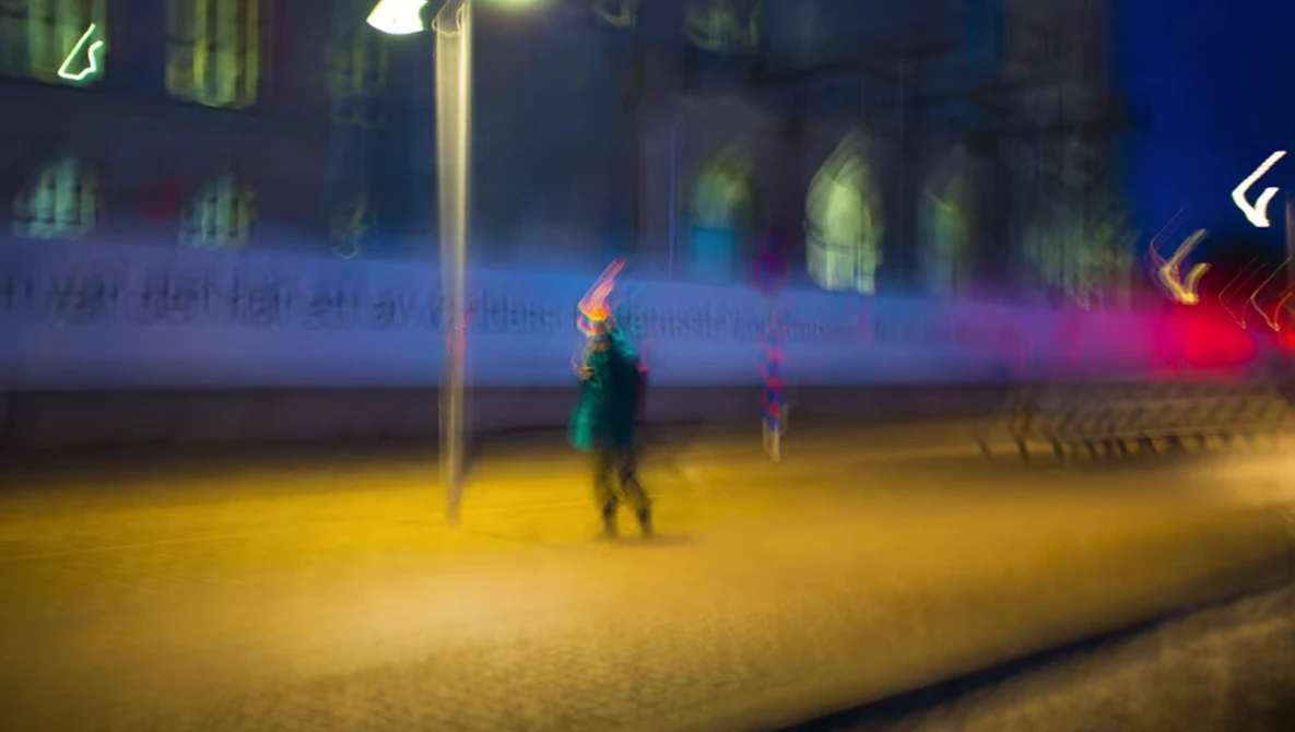 Blurred figure in green jacket standing in yellow-lit outdoor plaza at night with colorful neon signs in background.