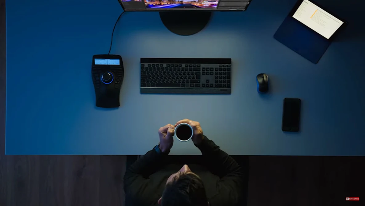 Overhead view of a person at a desk holding a coffee cup, surrounded by computer equipment on a blue surface.