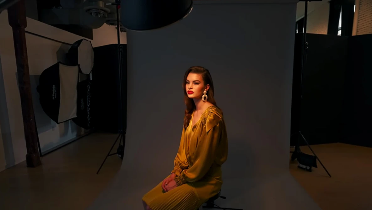 Woman in mustard-yellow dress seated in professional studio with multiple studio lights and reflectors.