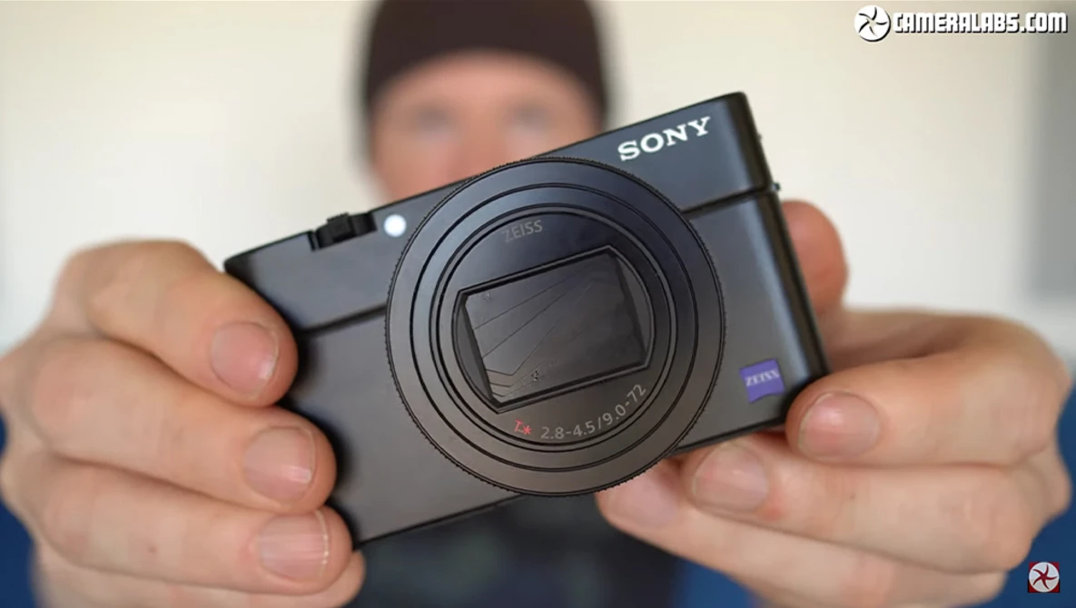 Hands holding a Sony compact camera with a zoom lens and blue Zeiss branding.