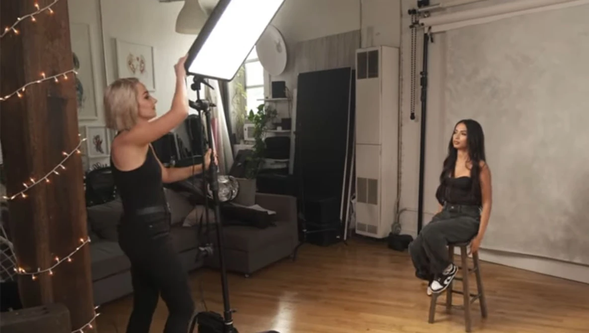 Studio portrait setup with female subject seated on stool while photographer adjusts overhead softbox lighting.