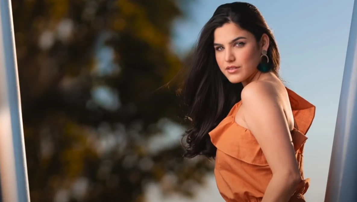 Woman with long dark hair wearing an orange sleeveless top, posed against a blurred natural background.