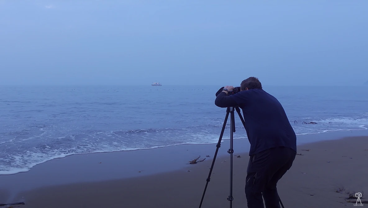 Photographer using tripod and telephoto lens on a beach at dusk, shooting toward the ocean horizon.