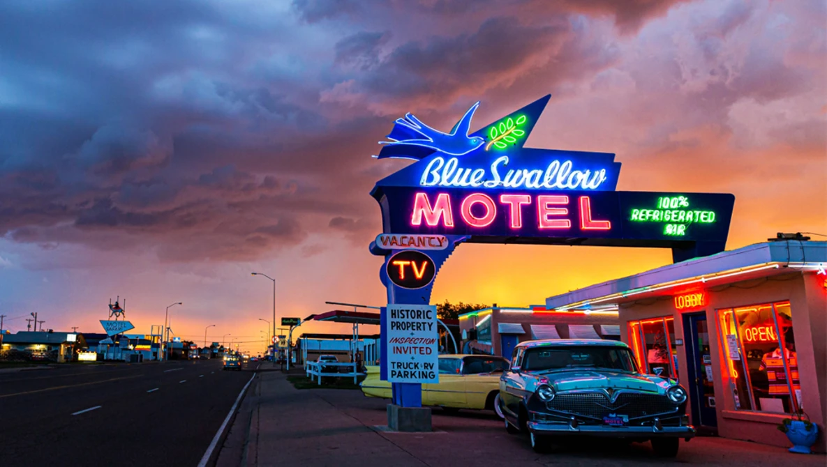 Blue Swallow Motel vintage neon sign glowing at dusk under a dramatic sunset sky with vintage cars parked outside.