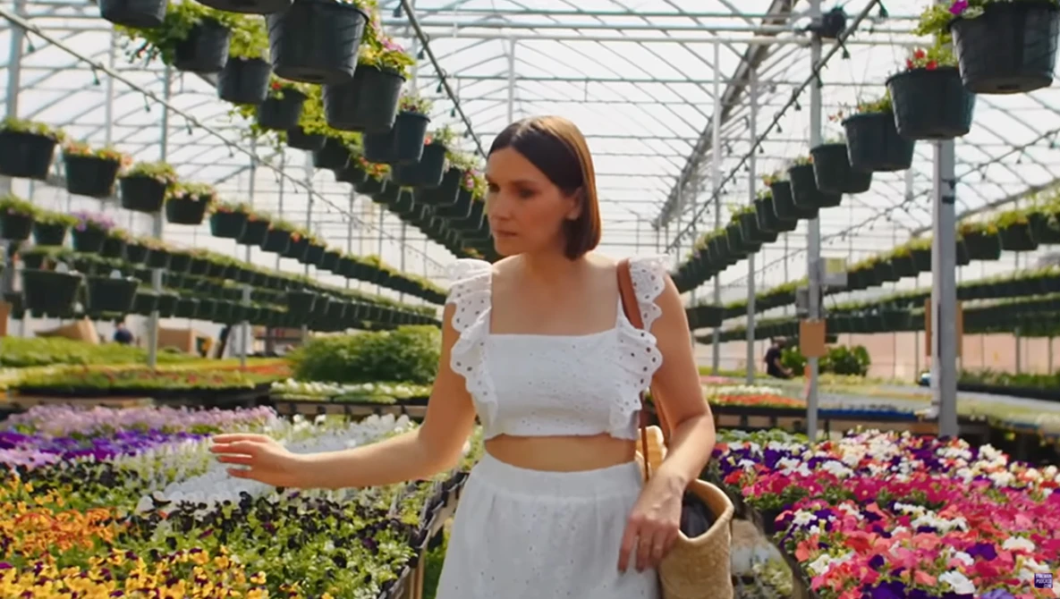Woman in white crop top and skirt standing in greenhouse surrounded by colorful flowering plants.