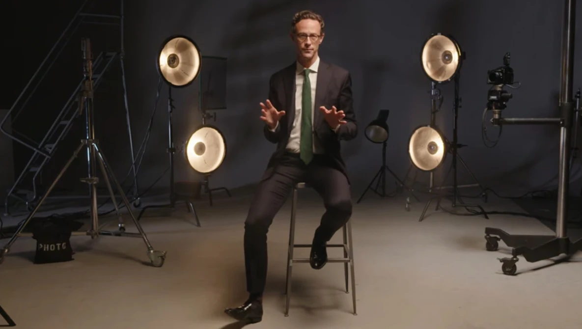 Man in business attire sitting on a stool in a professional studio setup with ring lights.