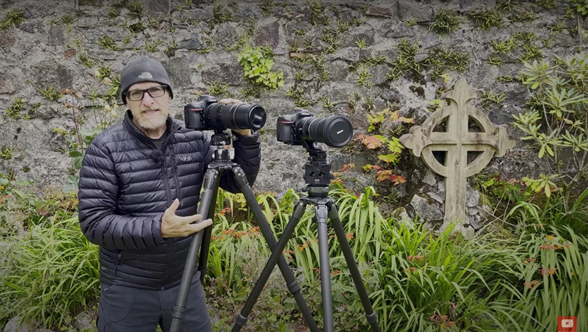 Photographer with telephoto lenses on tripods in front of moss-covered stone wall and cross.