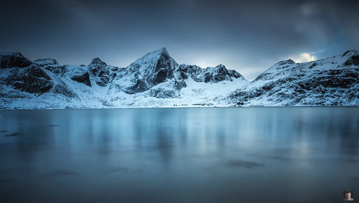 Snow-covered mountain peaks reflected in a still, partially frozen alpine lake at twilight.