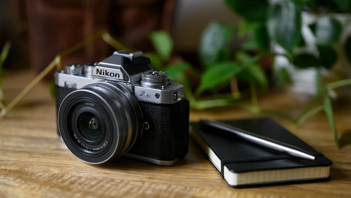 Nikon Z camera with attached lens on wooden desk beside closed notebook, indoor plants visible in background.