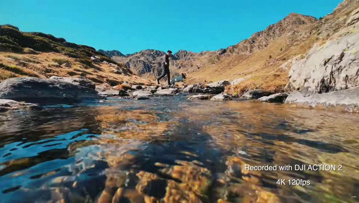 Shallow mountain stream flowing over rocky bottom with hikers standing in distant valley under clear sky.