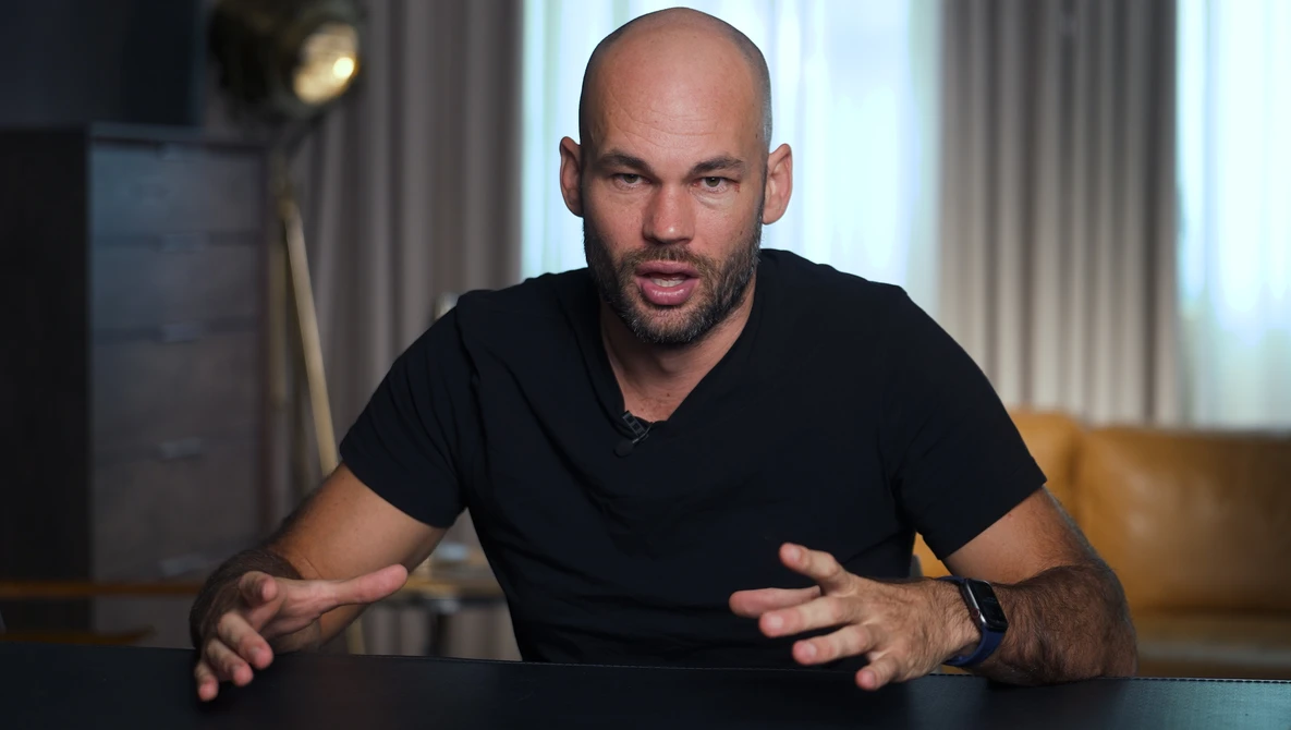 Man in black v-neck t-shirt gestures while speaking during an interview or presentation in a modern interior.