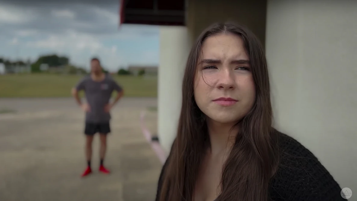 Young woman with long dark hair looking toward camera while person exercises in blurred background.