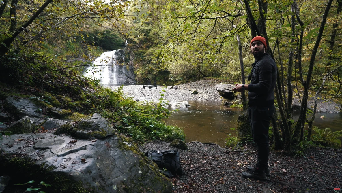 Photographer in dark clothing and orange beanie standing on rocky stream bank with waterfall visible in forested background.