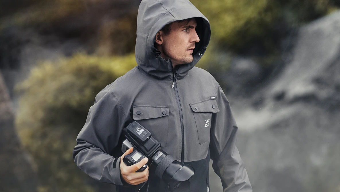 Photographer wearing gray technical jacket and hood while holding a telephoto lens outdoors.