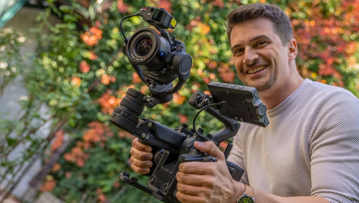 Filmmaker holding a cinema camera rig with mounted gimbal stabilizer against a flowering vine backdrop.