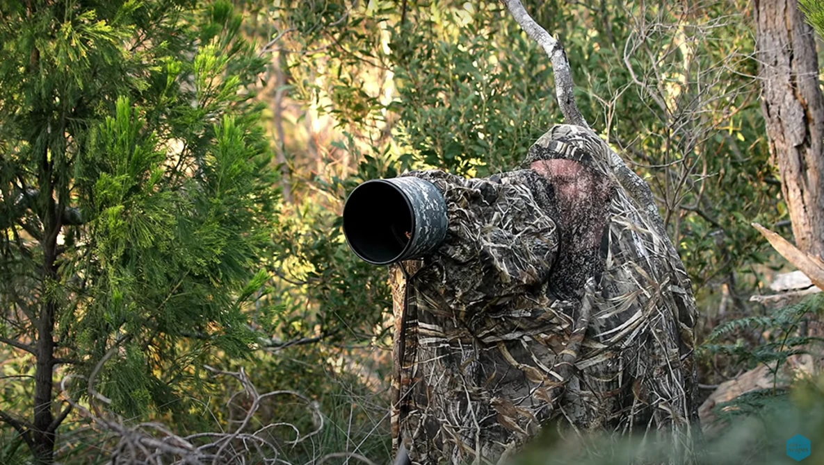Wildlife photographer in full ghillie suit aiming telephoto lens toward forest canopy.