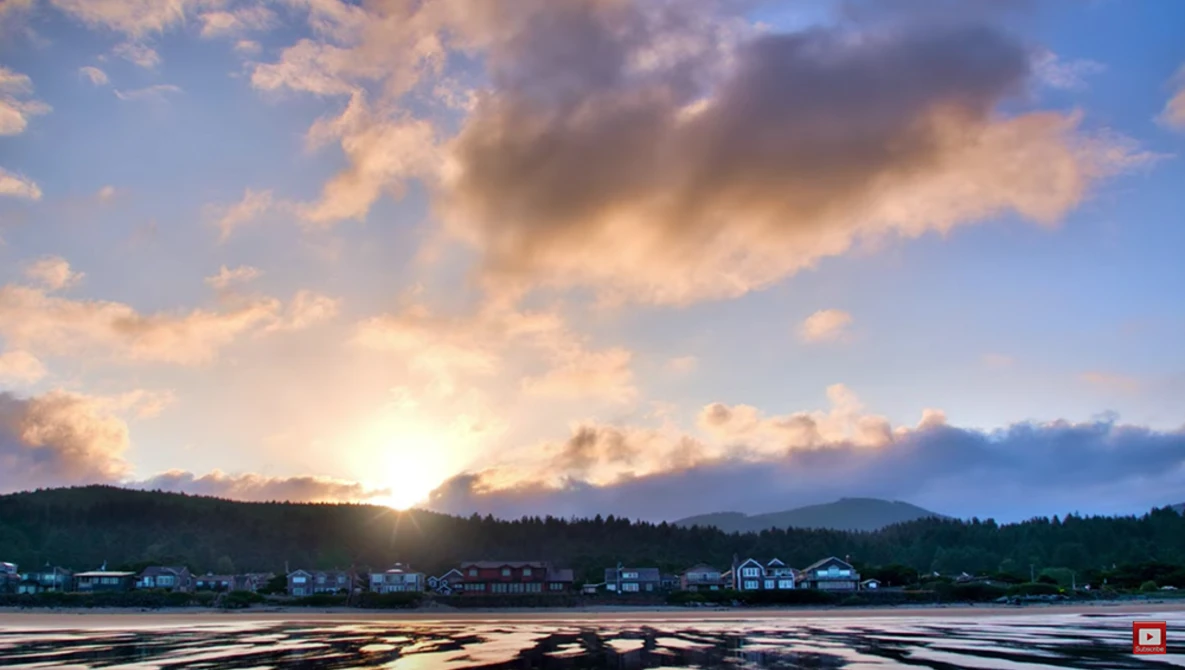 Sunset over a frozen lake with houses and forested hills silhouetted against golden clouds.