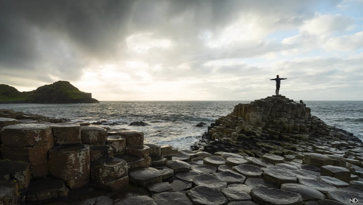 Figure standing on a rocky outcrop with a cross silhouetted against a dramatic cloudy seascape.