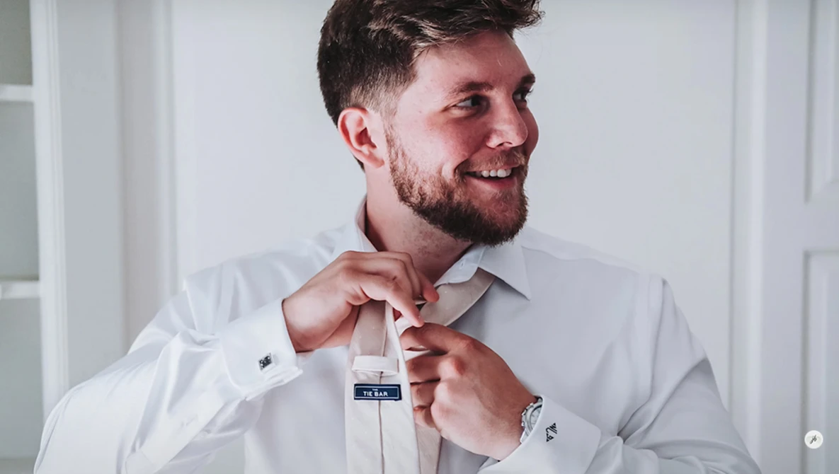 Man in white dress shirt adjusting tie with both hands while smiling.