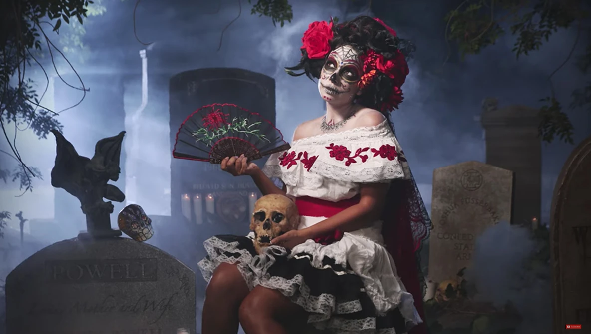 Woman in skull makeup and floral crown holding a hat in a cemetery at dusk.