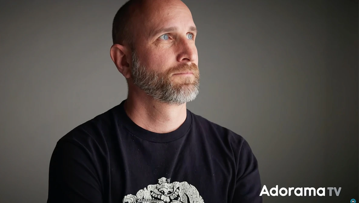 Man with beard wearing black t-shirt against neutral gray background, looking upward.
