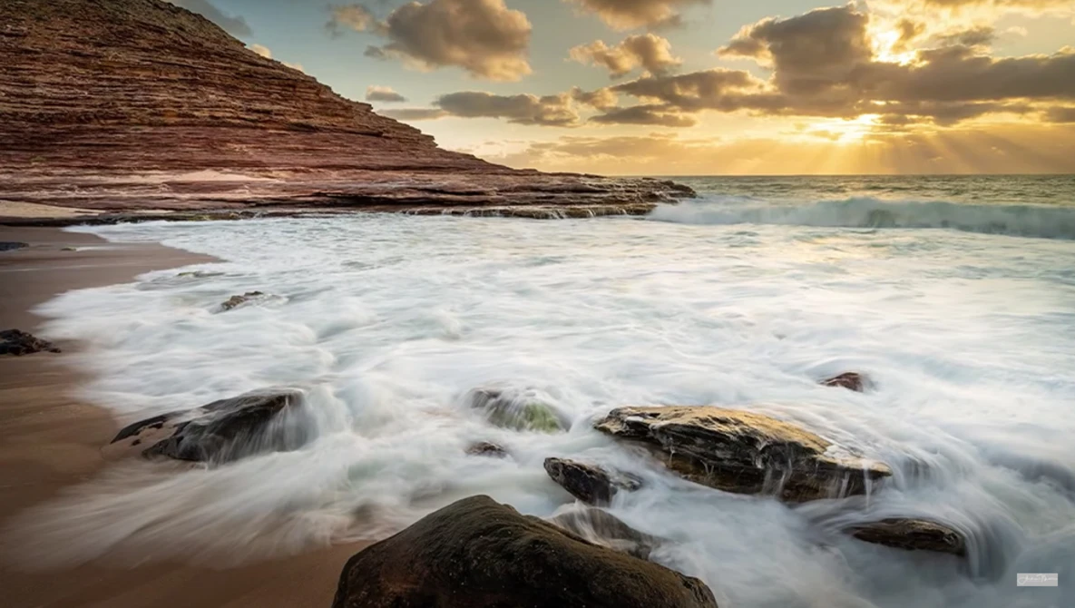 Waves roll onto a rocky beach at sunset, with layered cliff formations rising in the background.