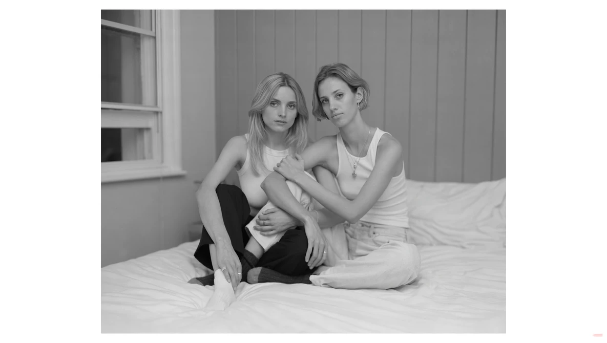 Two women sitting together on a bed, smiling at the camera in a black and white portrait.