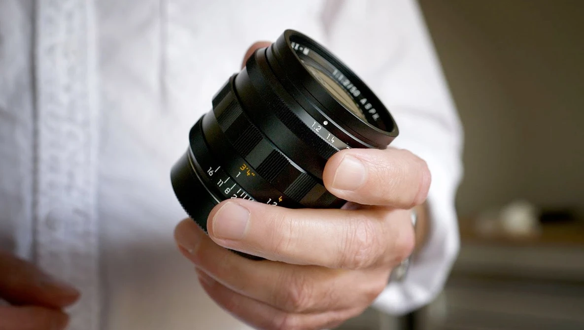 Hand holding a vintage manual focus lens with aperture ring and distance scale markings visible.