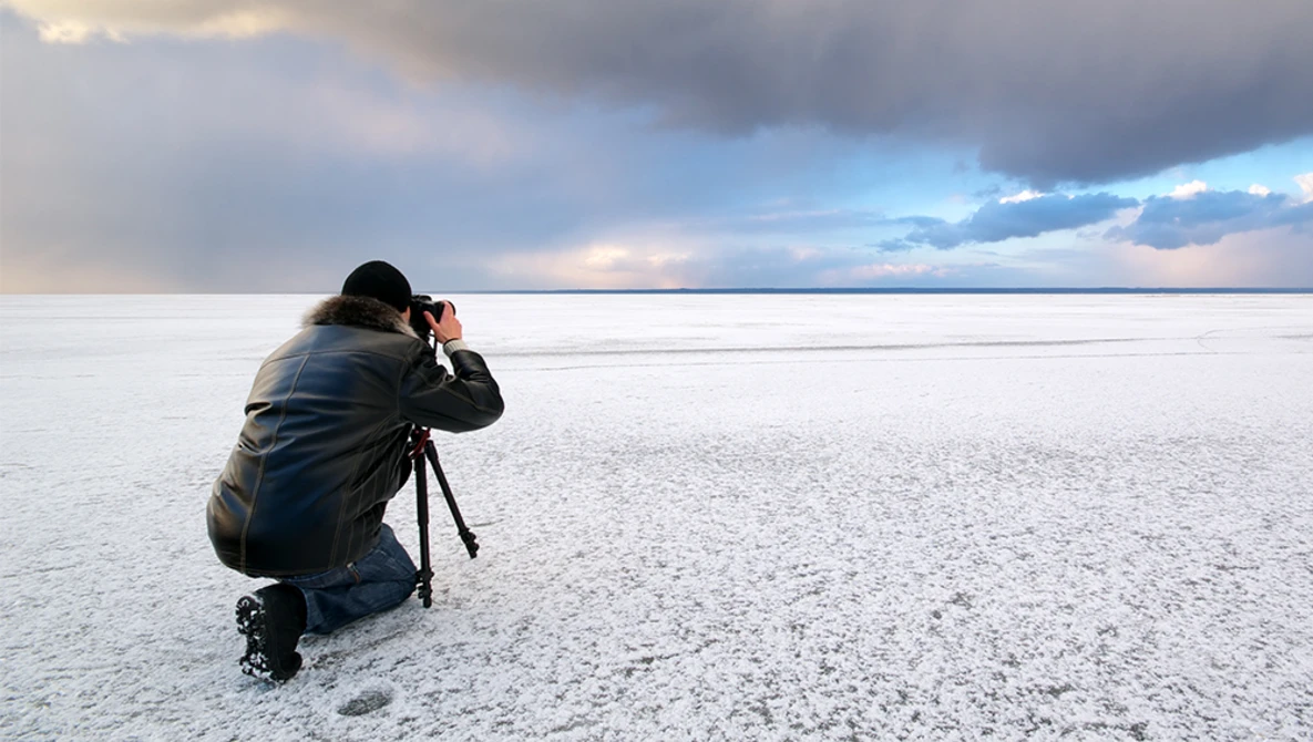 Photographer crouching on a salt flat, composing a shot with a telephoto lens during dramatic cloud-filled sunset.