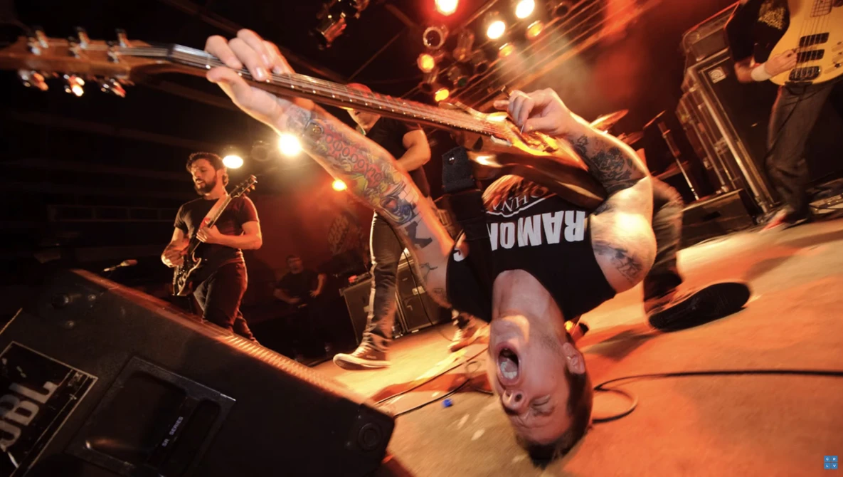 Musician performing a backflip while playing guitar on an illuminated stage.
