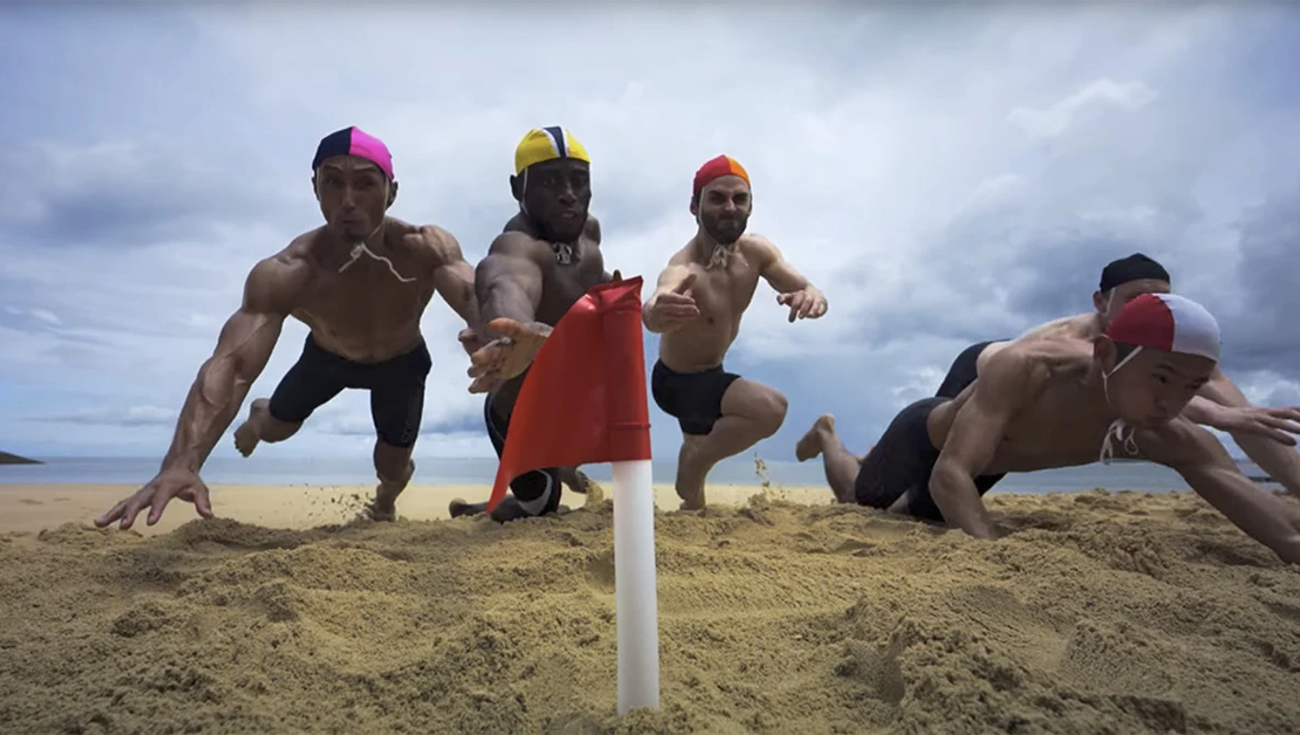 Five shirtless men in colored caps doing push-ups on sand with a white pole marker and cloudy sky behind them.