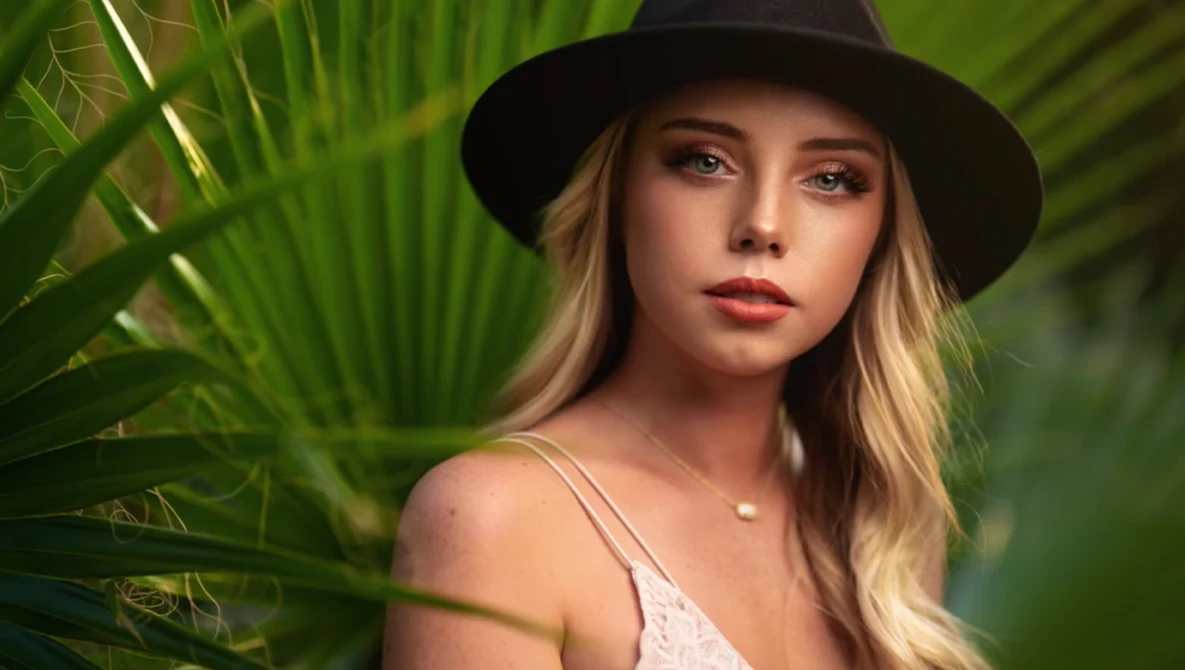 Woman wearing a black cowboy hat and white tank top, posed among green palm fronds.