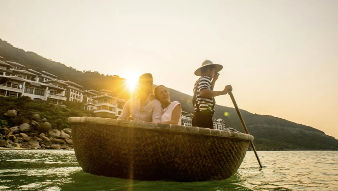 Family in a traditional woven boat on calm water during golden hour with hillside buildings in background.