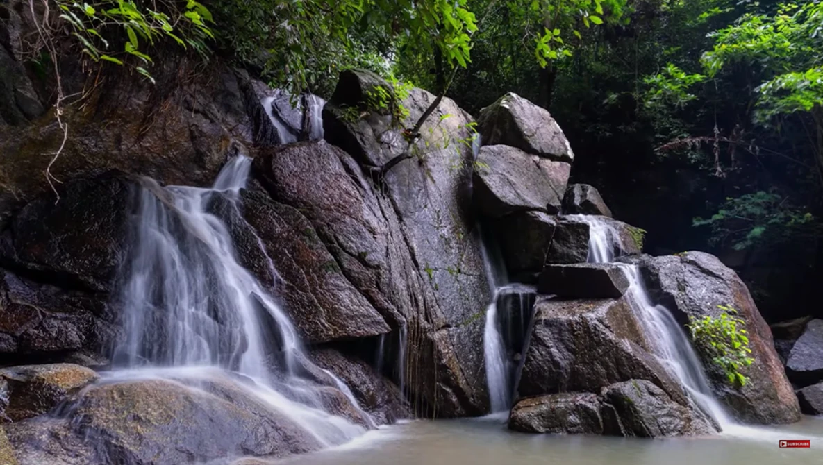 Multi-tiered waterfall cascading over large boulders surrounded by dense green forest.