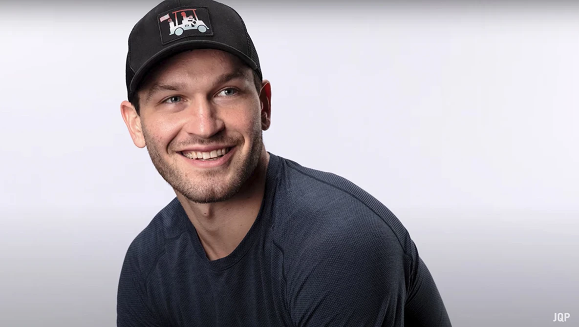 Smiling man wearing a black baseball cap and dark blue t-shirt against a neutral gray backdrop.