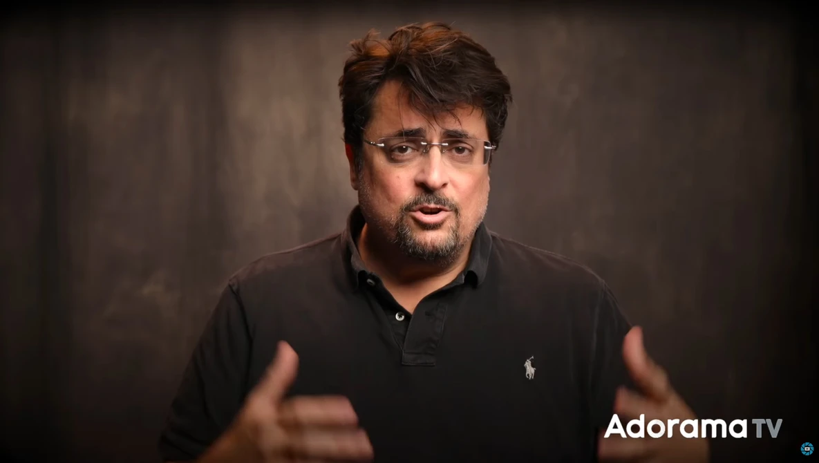 Man in black polo shirt gesturing while speaking against neutral backdrop.