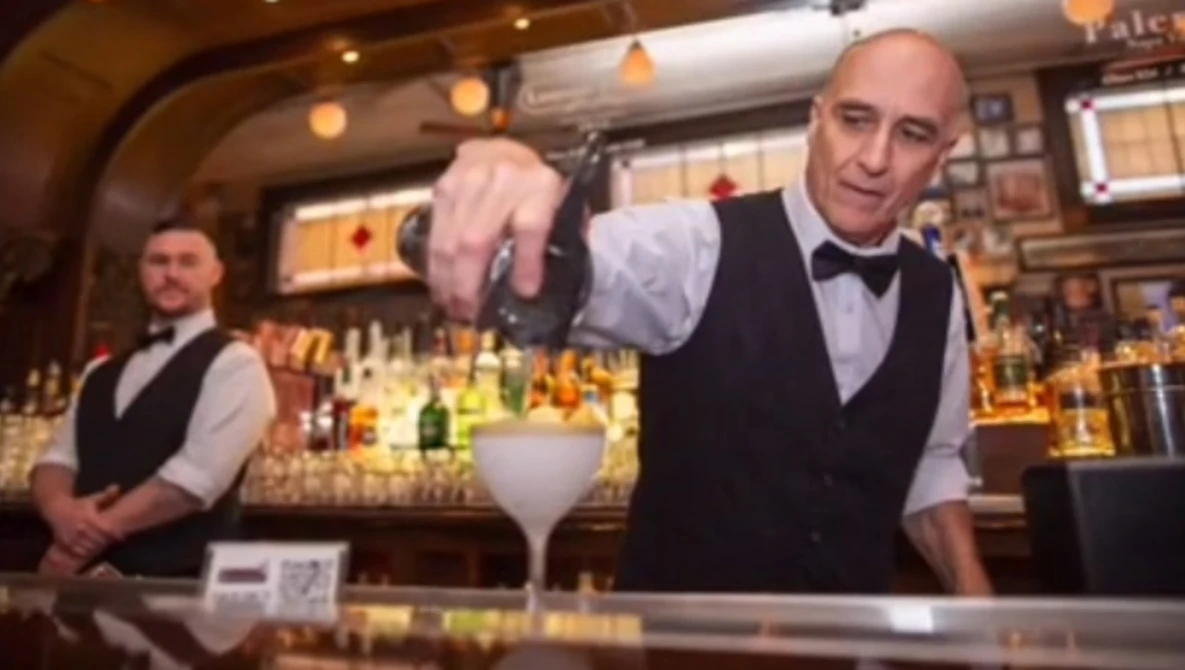 Bartender in formal attire pouring an ingredient into a cocktail glass behind an upscale bar counter.