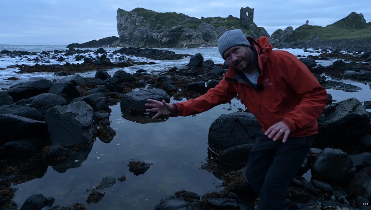 Photographer in red jacket reaching toward tide pool rocks during golden hour coastal shoot.
